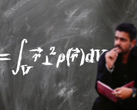 chalkboard in the background with a lengthy formula written on it. A blurred image of a man sitting in front of the board, book in hand, other hand at mouth/chin. Looking off into the distance in thought.